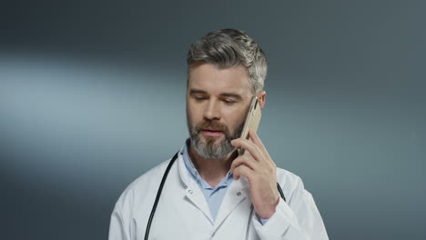 Caucasian-male-physician-speaking-on-the-mobile-phone-and-doing-consultation-by-connection-on-the-gray-wall-background.